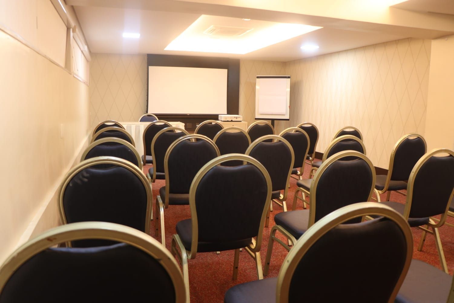 Sala de conferências vazia com fileiras de cadeiras pretas voltadas para uma tela de projeção e um flip chart.
