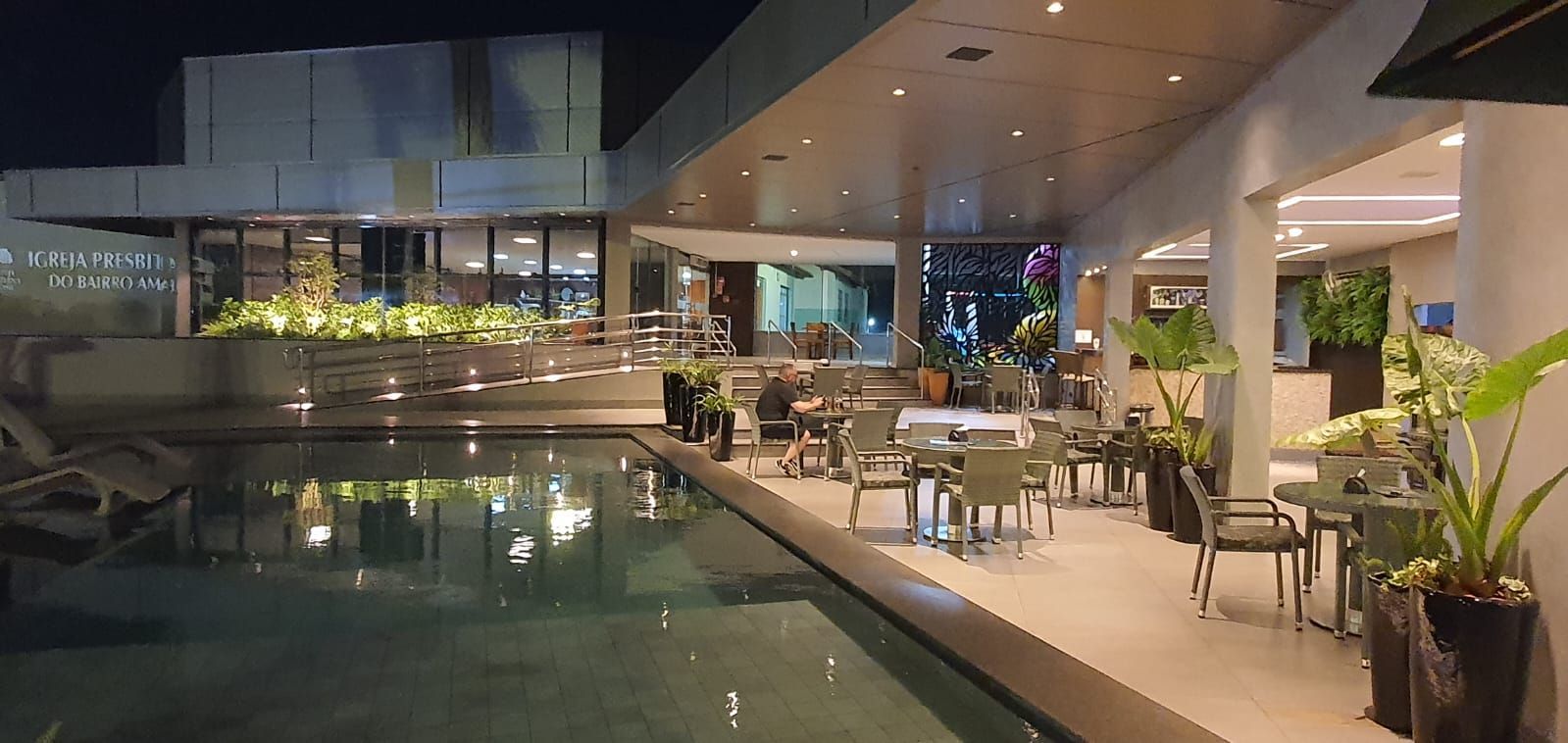 Terraço moderno de restaurante ao ar livre junto a uma piscina iluminada à noite, com mesas, cadeiras e vasos de plantas.
