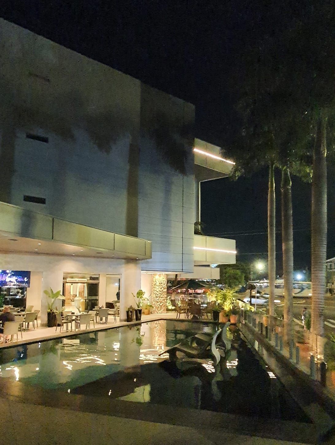 Pátio moderno do hotel à noite com piscina iluminada, palmeiras e espreguiçadeiras.