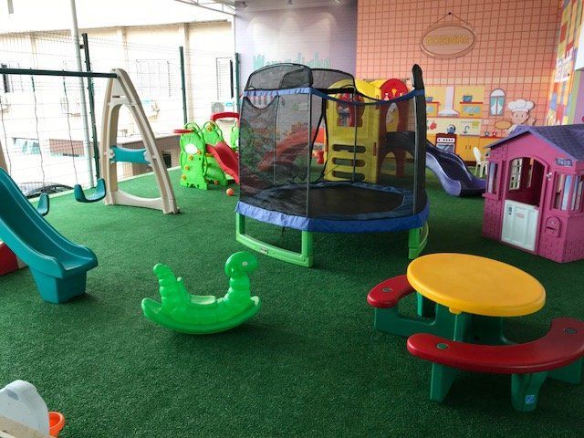 Parque infantil coberto com grama sintética, escorregador, cama elástica, mesa de piquenique e casinha de brincar sob luzes brilhantes.