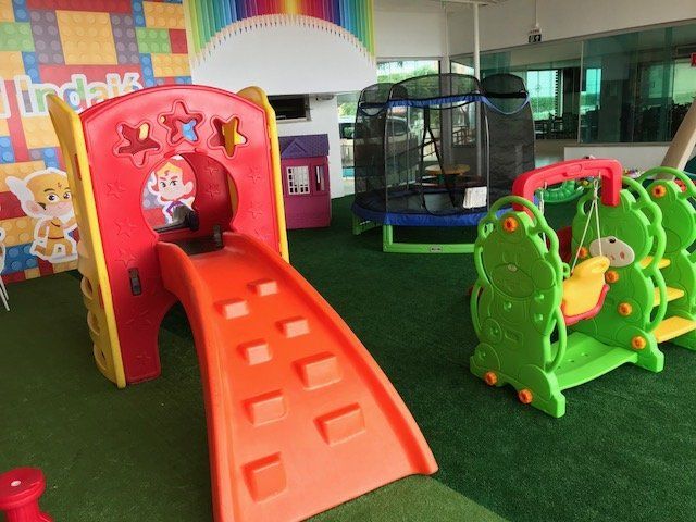 Área de recreação infantil com escorregador vermelho e brinquedos de mola verdes em grama artificial.