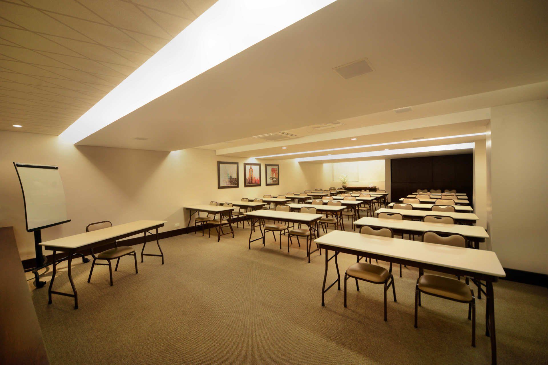 Sala de aula vazia com fileiras de mesas e cadeiras, tela de projeção e iluminação quente no teto.