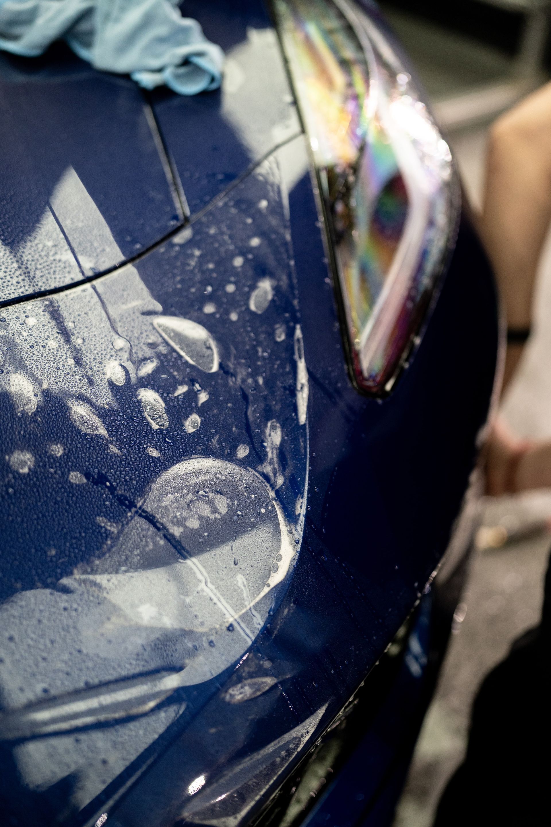 Close-up of a blue car covered in water droplets, with a visible headlight and hand at the edge.