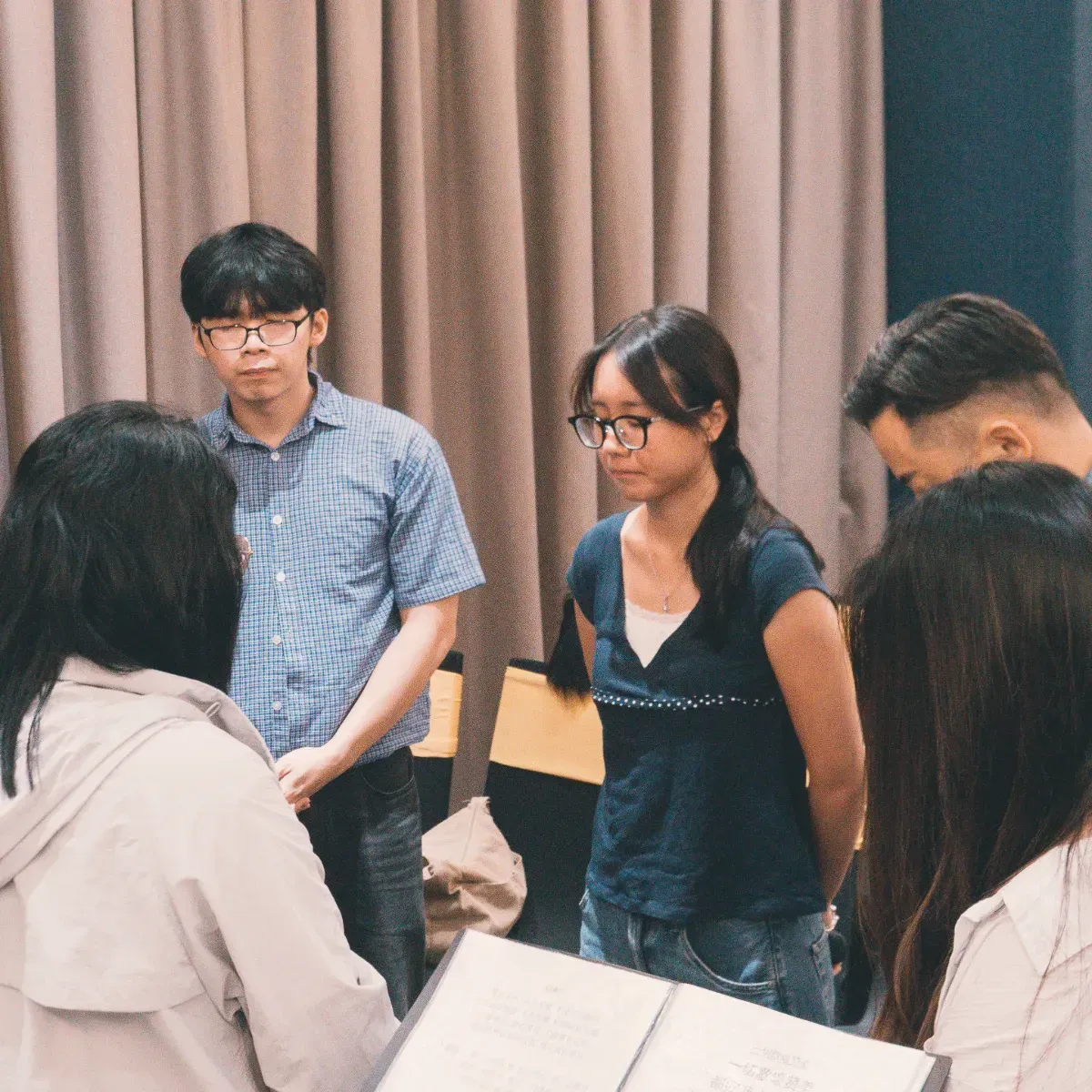 Small group standing in a room, talking near a curtain backdrop and chairs.