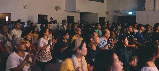 Audience seated in a dim auditorium watching a presentation, many looking toward the front screen
