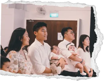People seated indoors holding babies at a ceremony, watching something ahead together.