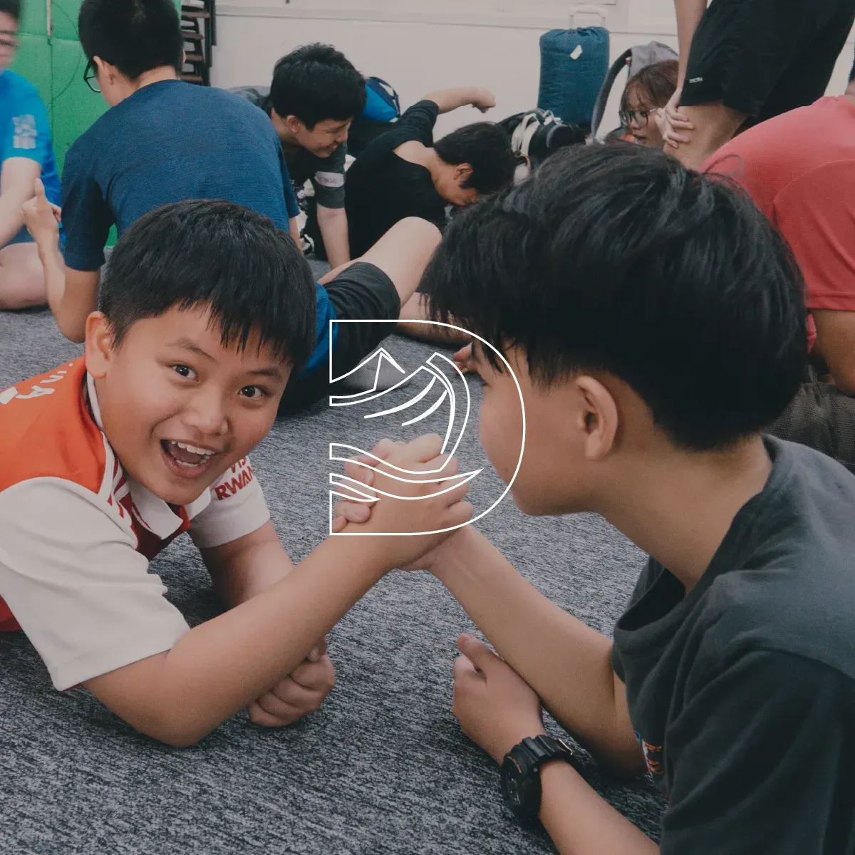 Two boys arm-wrestling on a carpeted floor while others sit nearby, one smiling at the camera