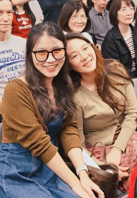 Two smiling women sit close together indoors, wearing glasses and casual sweaters.