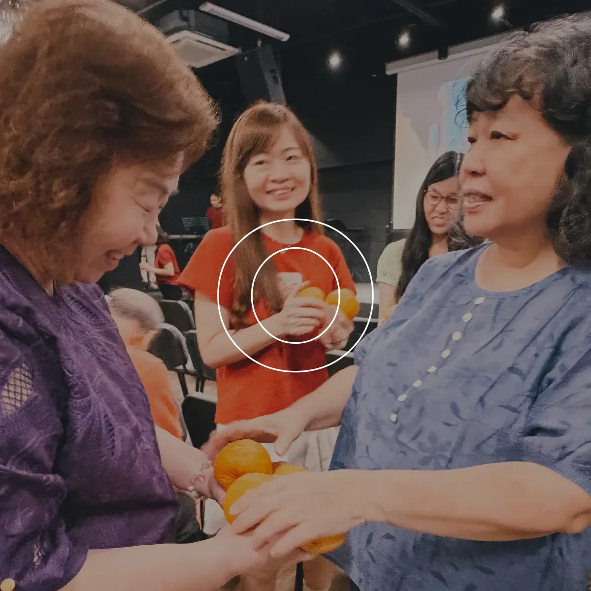 People exchanging oranges at a lively indoor gathering, with smiling attendees in the background