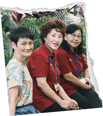 Three smiling people in red shirts sit together outdoors among green plants.