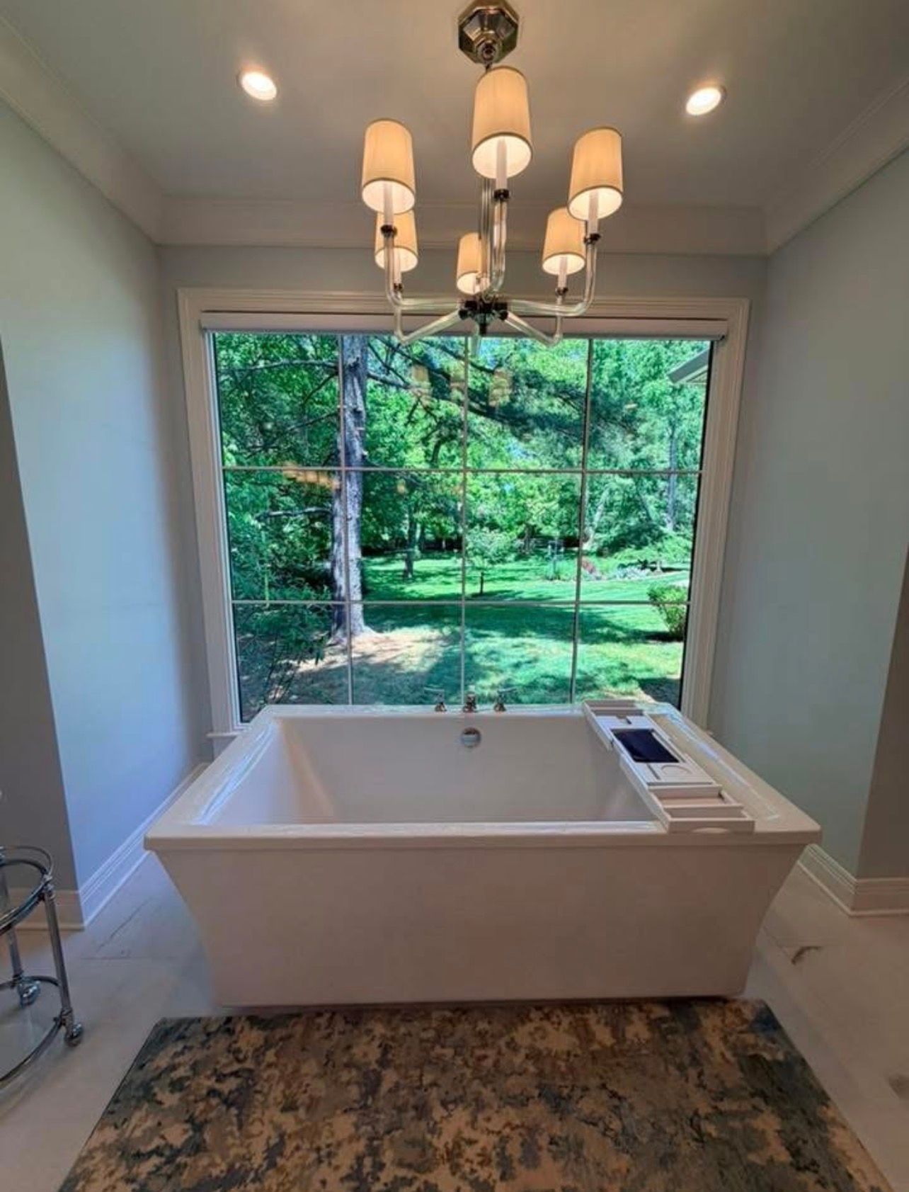 Bright bathroom with a white soaking tub beneath a chandelier and a large window overlooking greenery