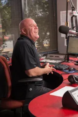 Matt Allen laughs while sitting at a desk in a radio studio with audio equipment, a microphone, and a computer monitor.