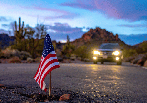 American flag on a desert roadside with headlights approaching against a mountain sunset backdrop.