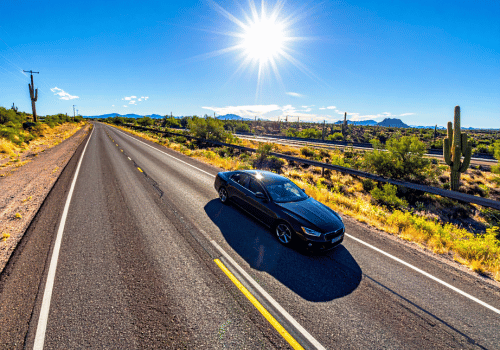 Black sedan driving along a sunlit desert highway surrounded by cacti, open landscape, and clear blue skies.
