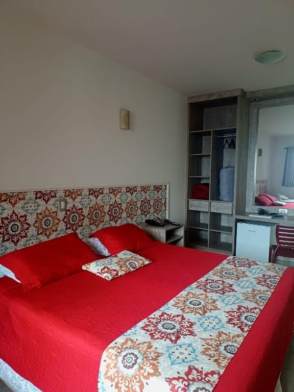 Cama vermelha com peseira e cabeceira estampadas em um quarto iluminado com guarda-roupa espelhado.