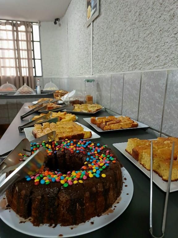 Bolo de chocolate e diversas sobremesas dispostas em uma longa mesa de bufê em uma sala com piso de azulejo.