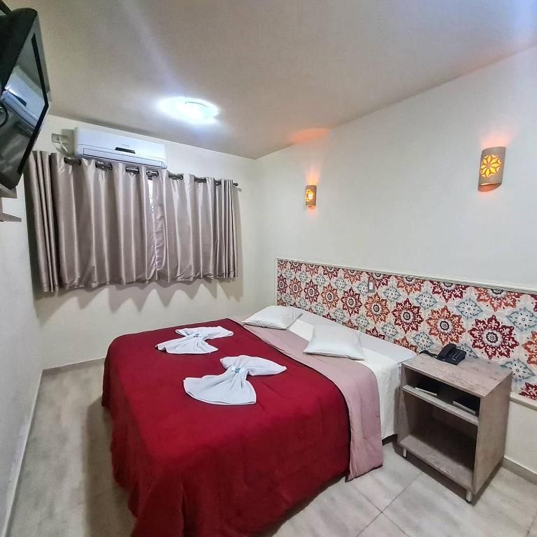 Quarto de hotel pequeno com colcha vermelha, toalhas brancas, cabeceira estampada, cortinas e apliques de parede.