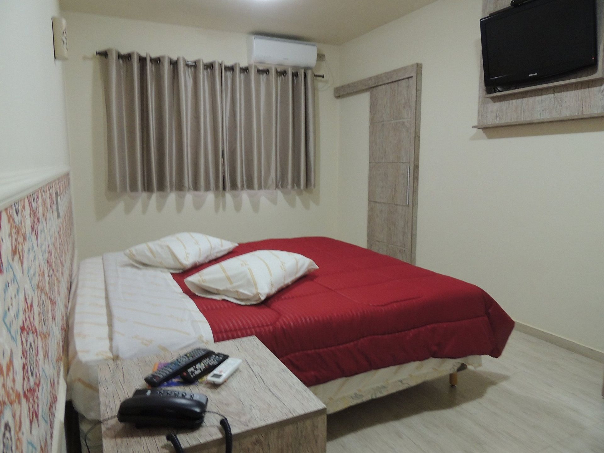 Quarto com cama desarrumada, cobertor vermelho, cortinas, mesa de cabeceira e TV fixada na parede.