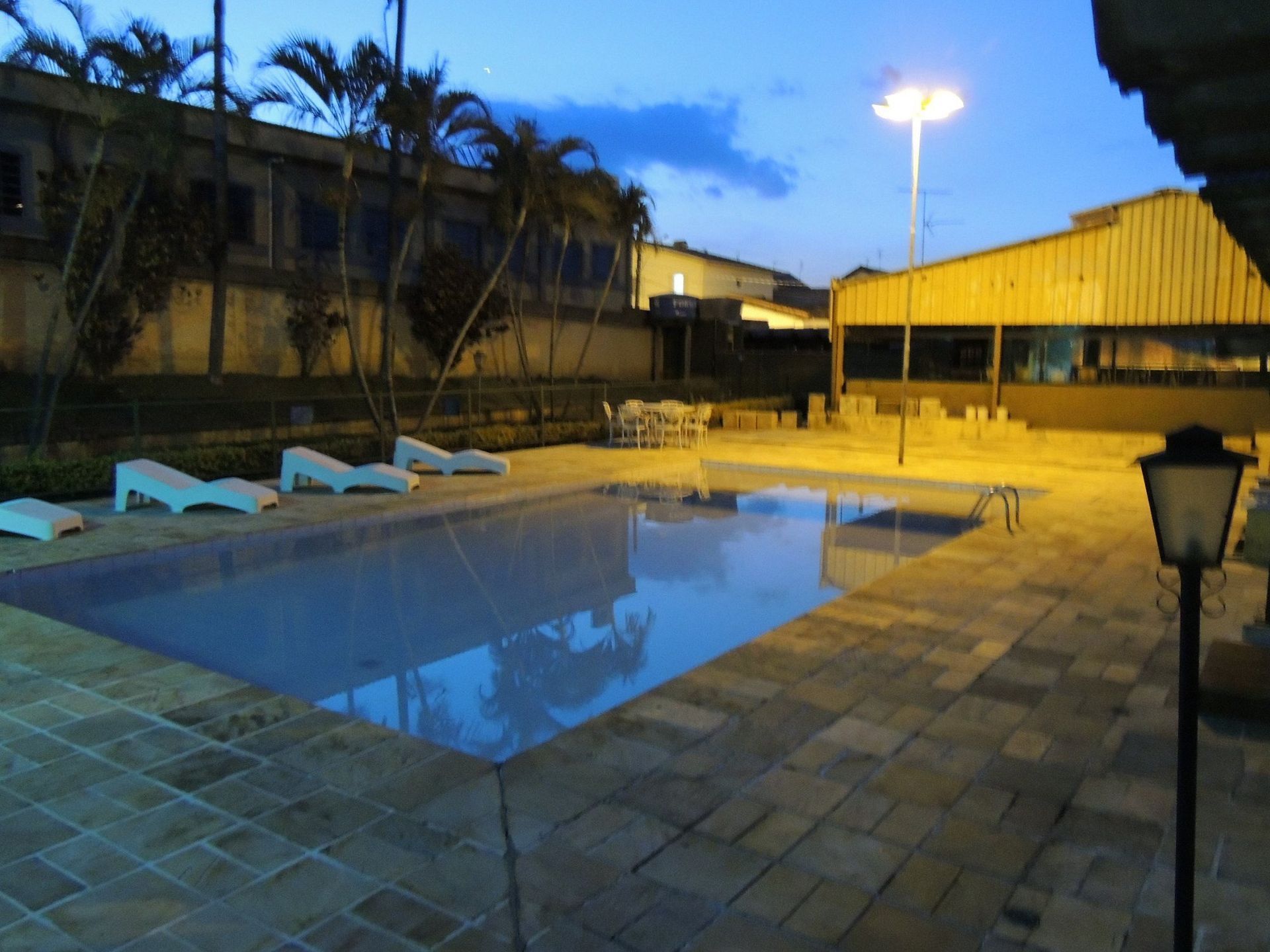 Piscina ao ar livre ao entardecer com espreguiçadeiras azuis, ladrilhos de pátio e reflexos dos edifícios iluminados na água.
