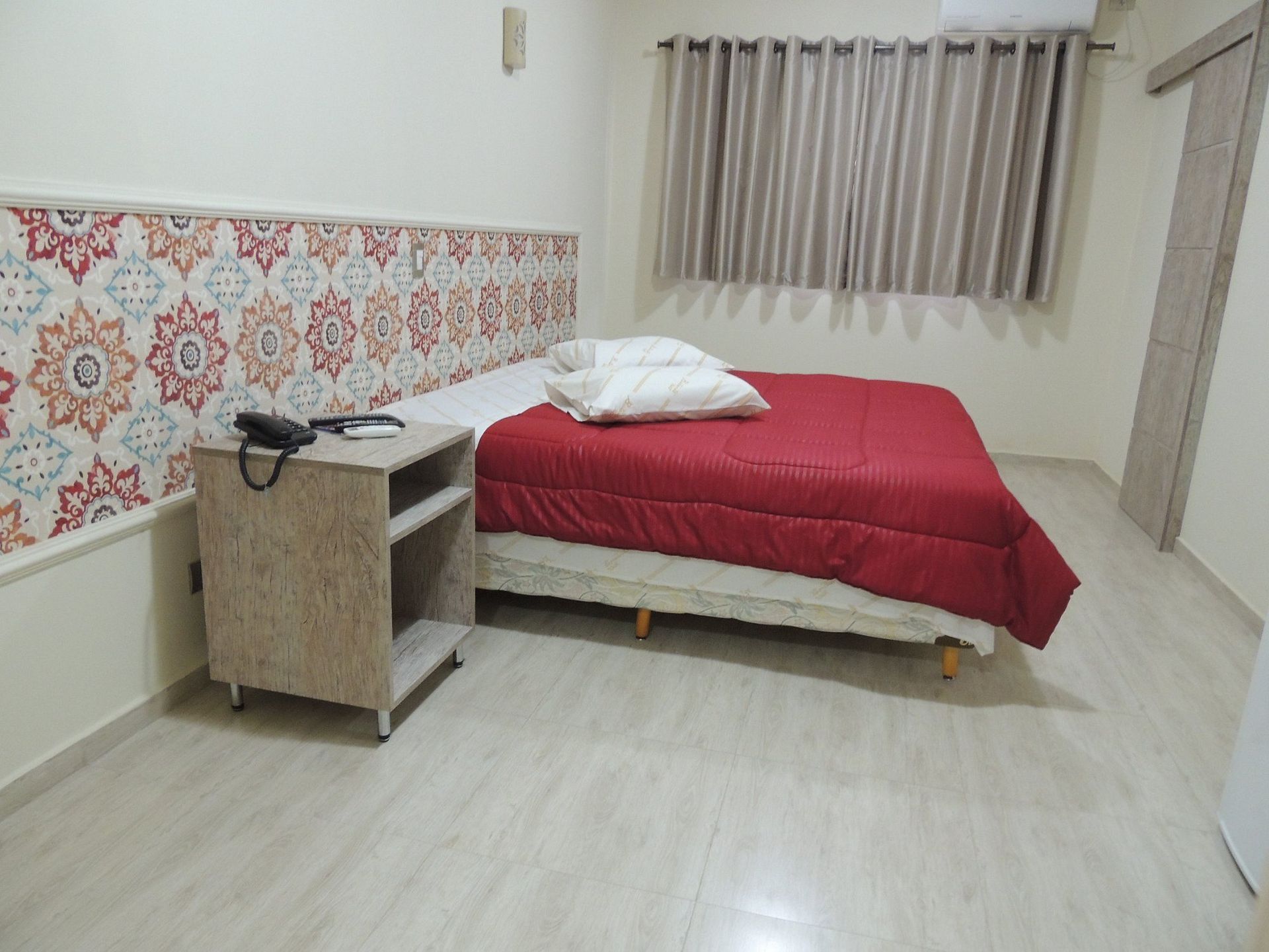 Quarto pequeno com colcha vermelha, travesseiros, mesa de cabeceira, faixa decorativa na parede e cortinas.
