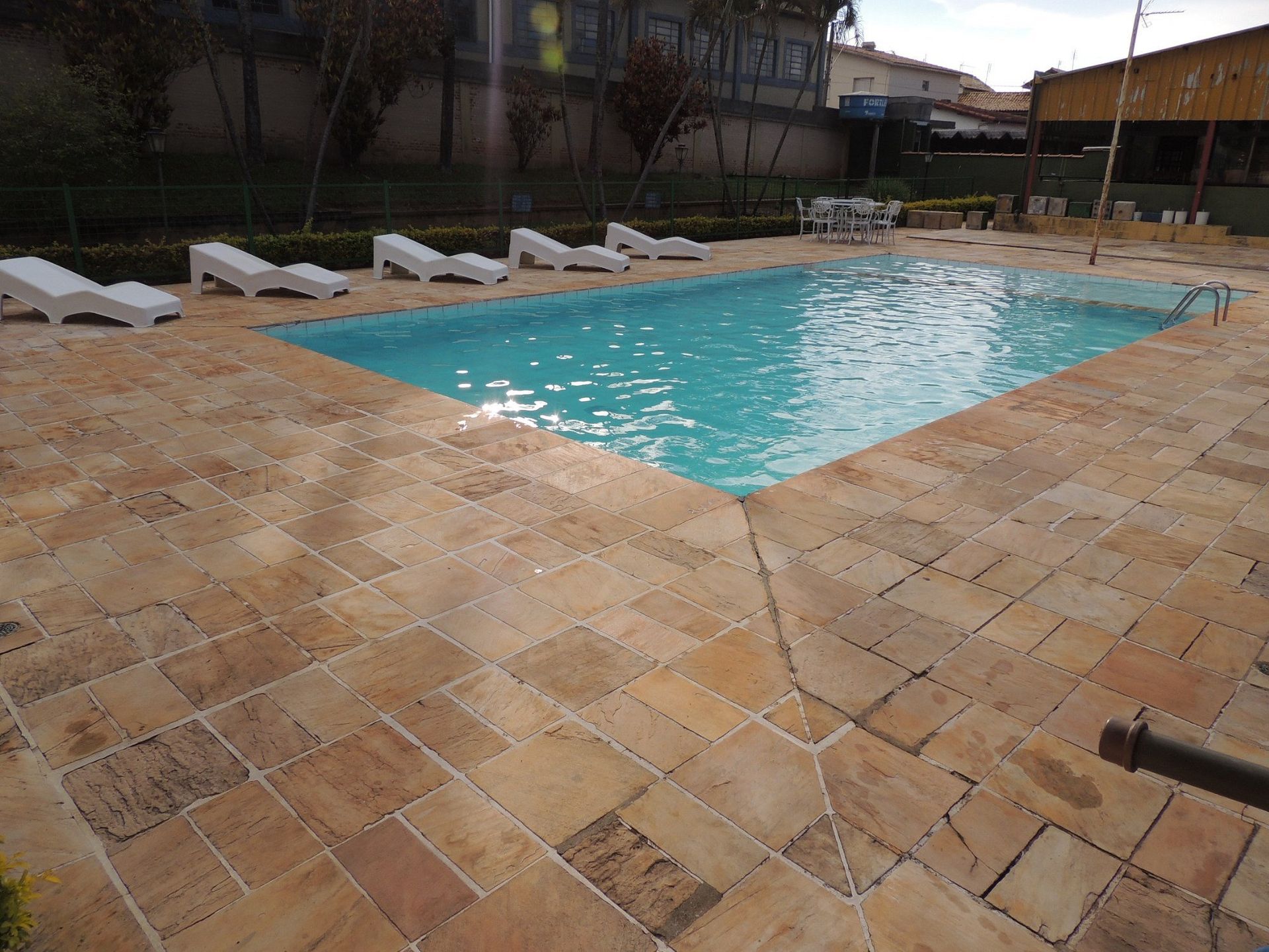 Piscina exterior com água turquesa, rodeada por ladrilhos de pedra cor de areia e espreguiçadeiras brancas.