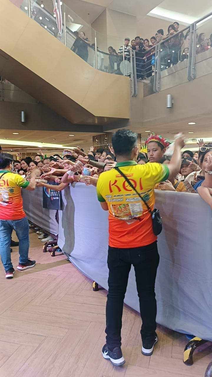 A man wearing a yellow shirt that says koi is standing in front of a crowd of people.