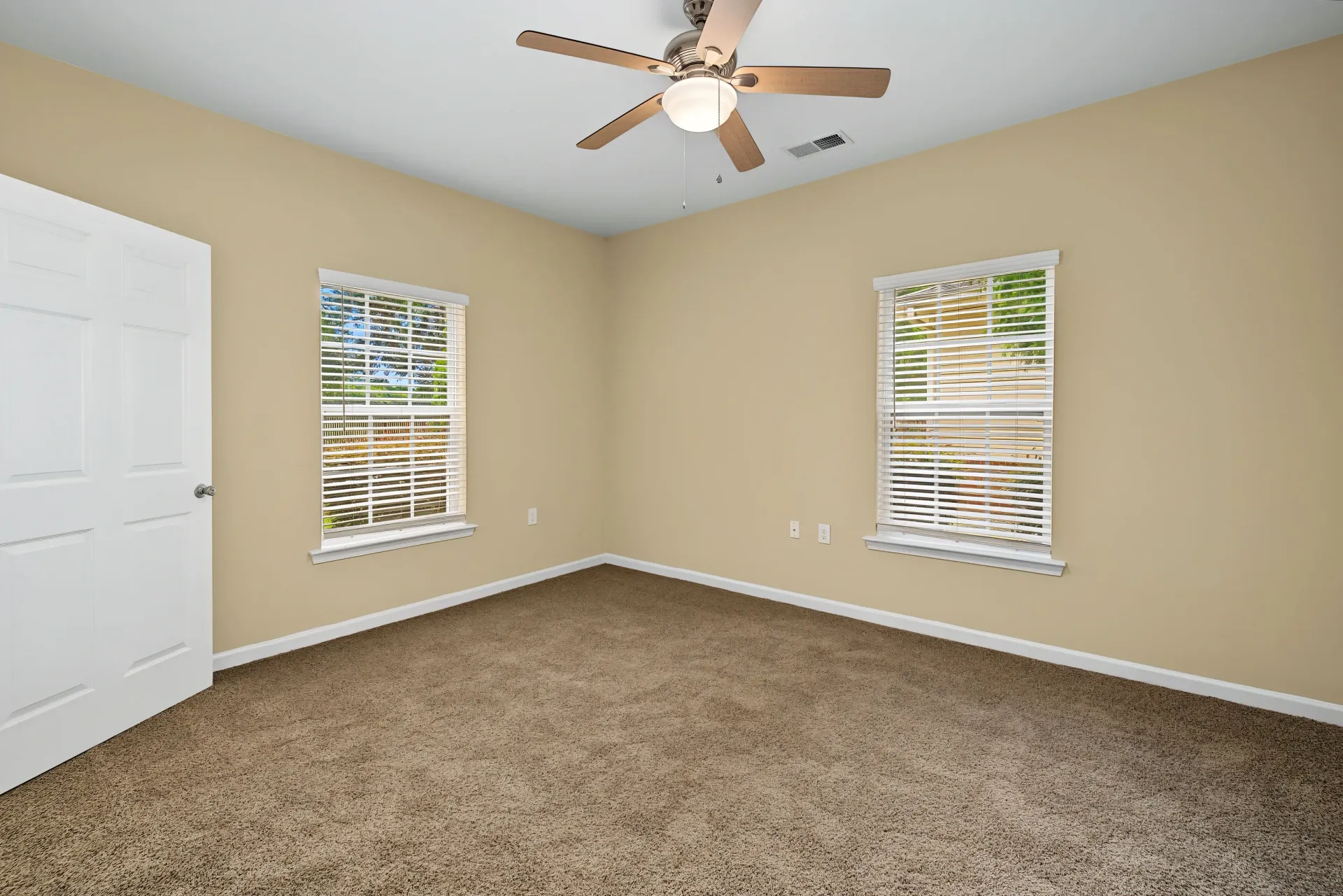 Empty bedroom with beige walls, brown carpet, a white door, a ceiling fan, and two windows with blinds.