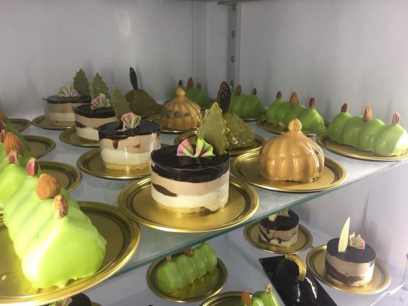 Un assortimento di pasticcini gourmet, tra cui torte al cioccolato a strati e dessert con glassa verde, disposti su scaffali espositivi.