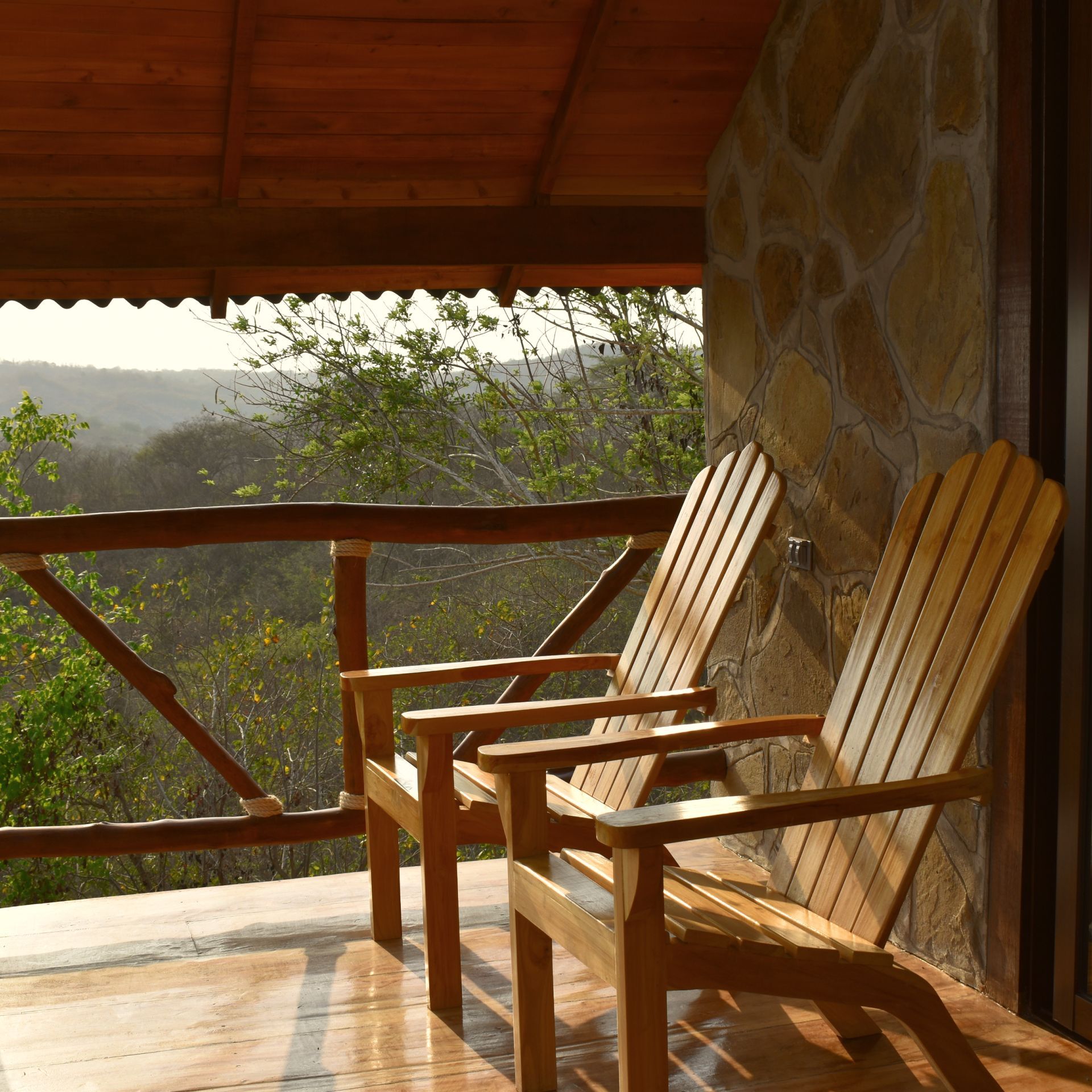 Dos sillas de madera en un balcón con vistas a un paisaje, muro de piedra a la derecha.
