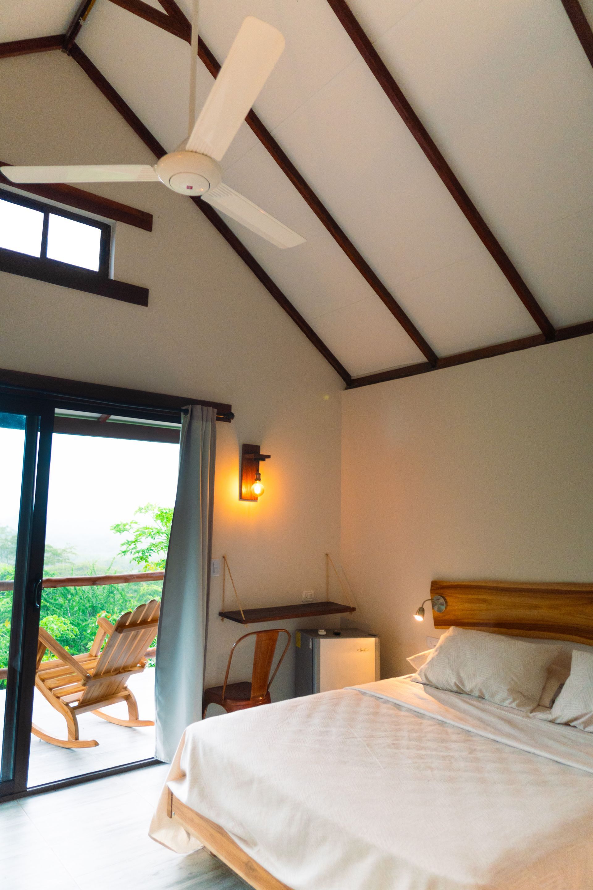 Dormitorio con cama, balcón y ventilador de techo. Paredes blancas y luminosas, molduras de madera.