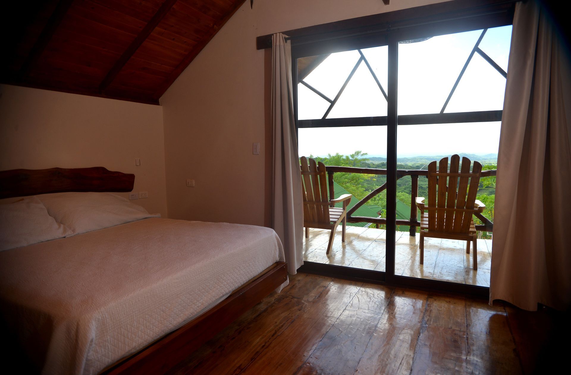 Dormitorio con cama, balcón con sillas, vista a los árboles y vista lejana.