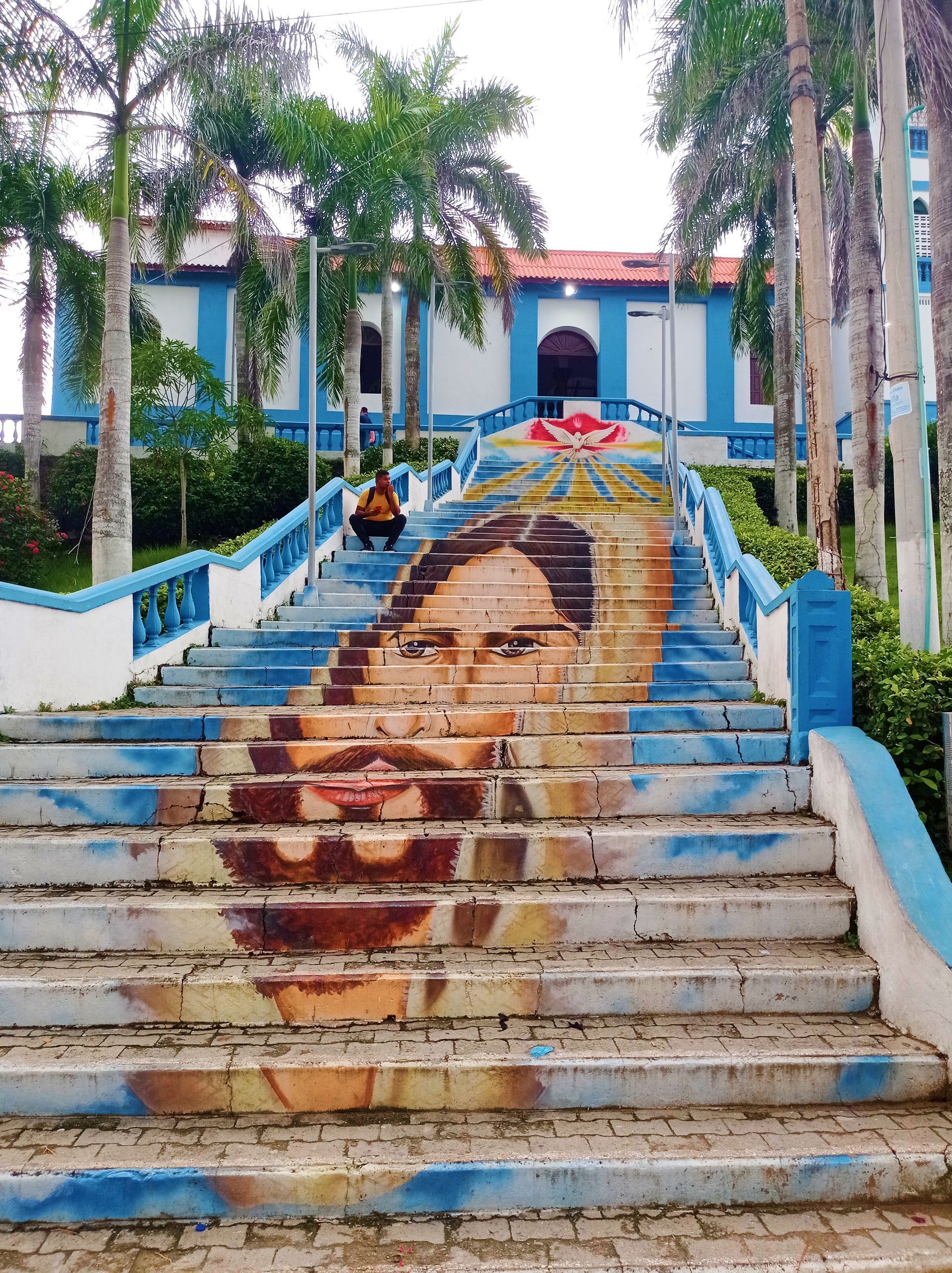Escalera pintada con un rostro de mujer que conduce a un edificio azul y blanco, con una persona sentada cerca de la parte superior.