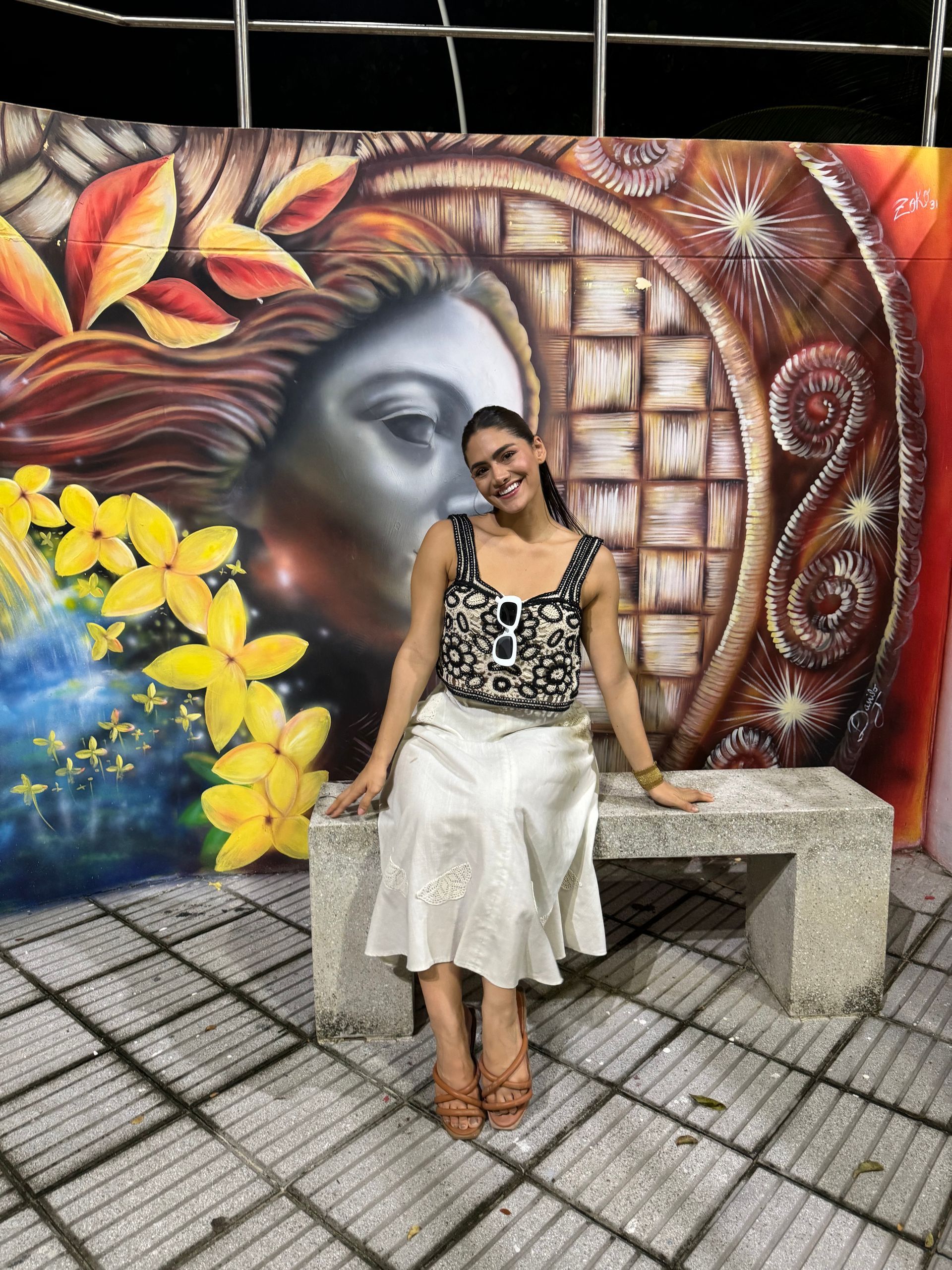 Mujer sentada en un banco de piedra frente a un mural colorido, sonriendo.