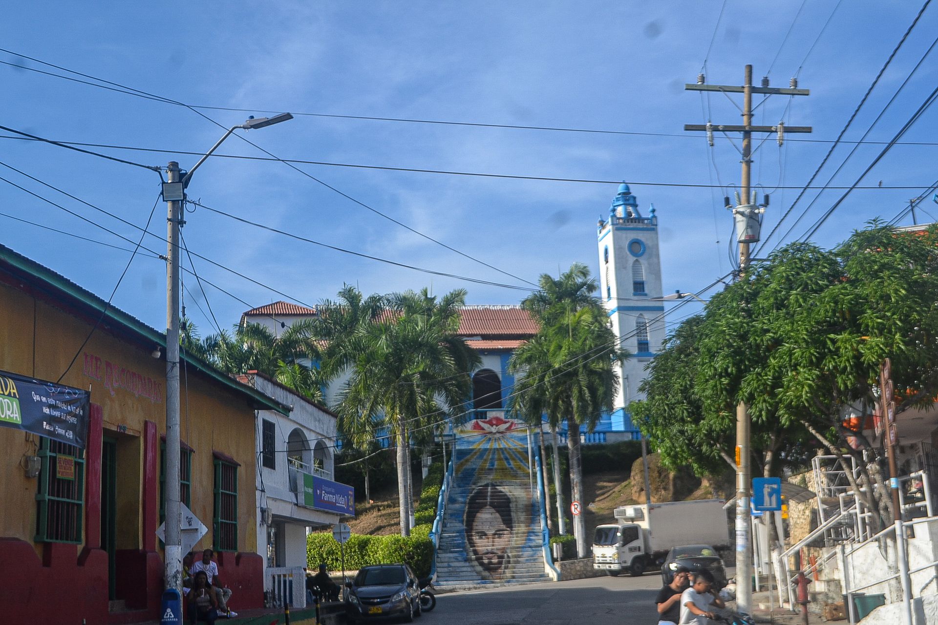 Vista de la calle de una iglesia con una torre azul y blanca, palmeras y un mural en los escalones.