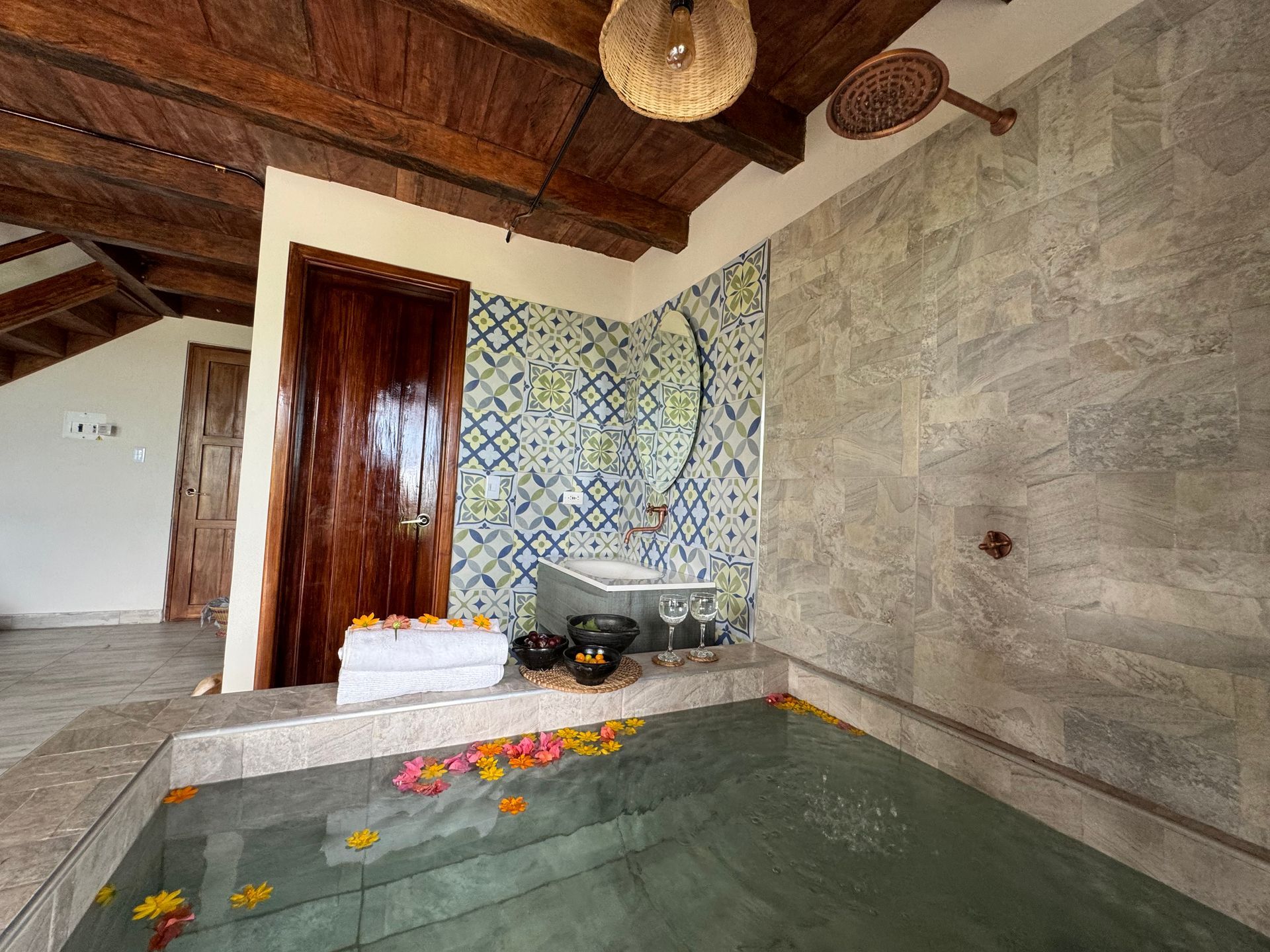 Baño con paredes de piedra, techo de madera y bañera hundida con pétalos de flores.