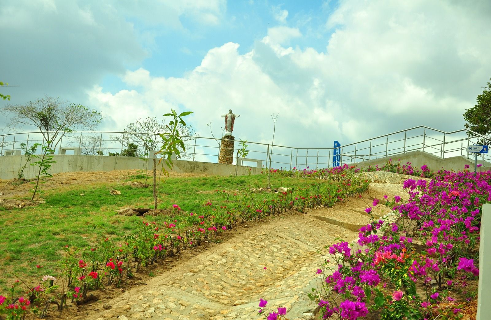 Estatua de Cristo en una colina, con vistas a un jardín paisajístico con flores rosas y rojas, cielo azul arriba.