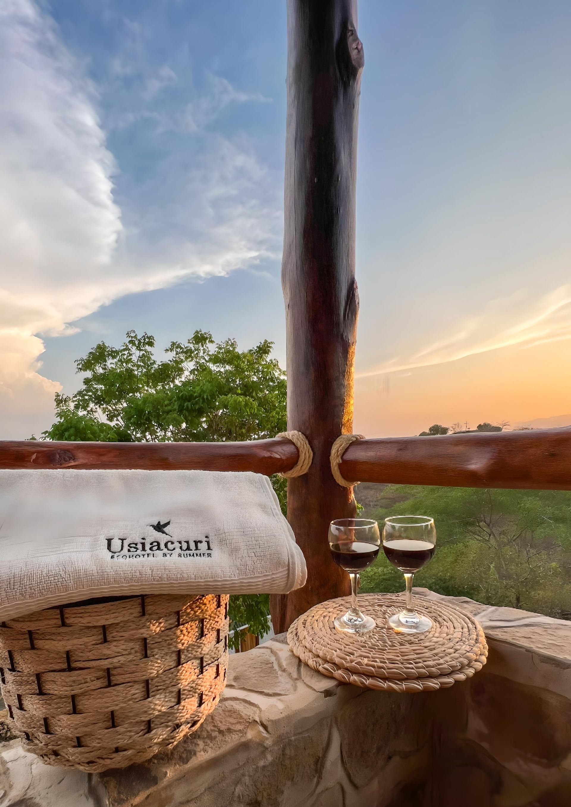 Cesta de mimbre con una toalla y dos copas de vino en una terraza con vistas a una puesta de sol.