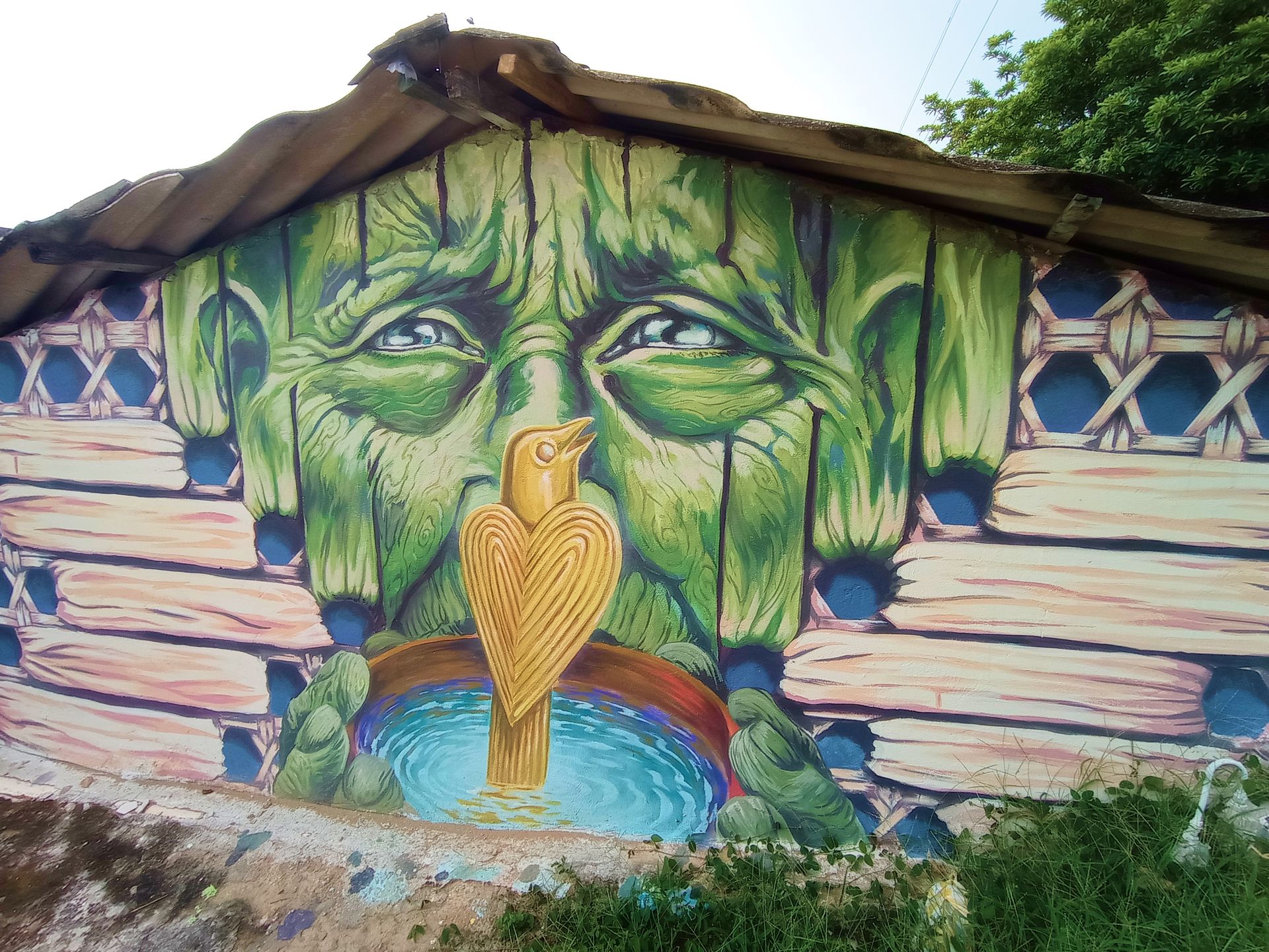 Mural de una figura de cara verde con un pájaro y un cuenco; situado sobre una pared de madera con techo de paja.