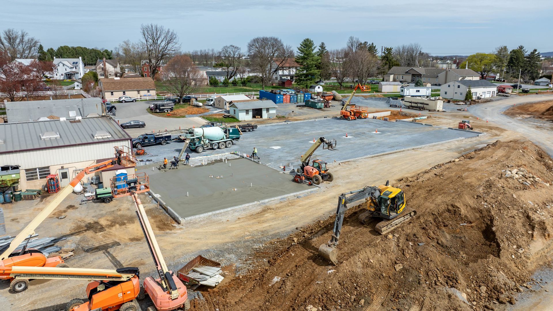 Construction site with excavators leveling a large dirt lot beside houses and trees