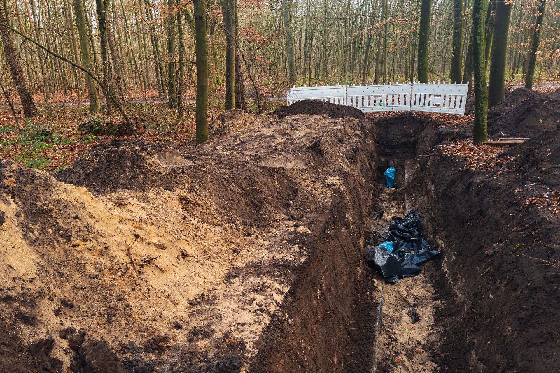 Deep muddy trench in a wooded area with an excavator and white fence in the background Deep muddy trench in a wooded area with an excavator and white fence in the background