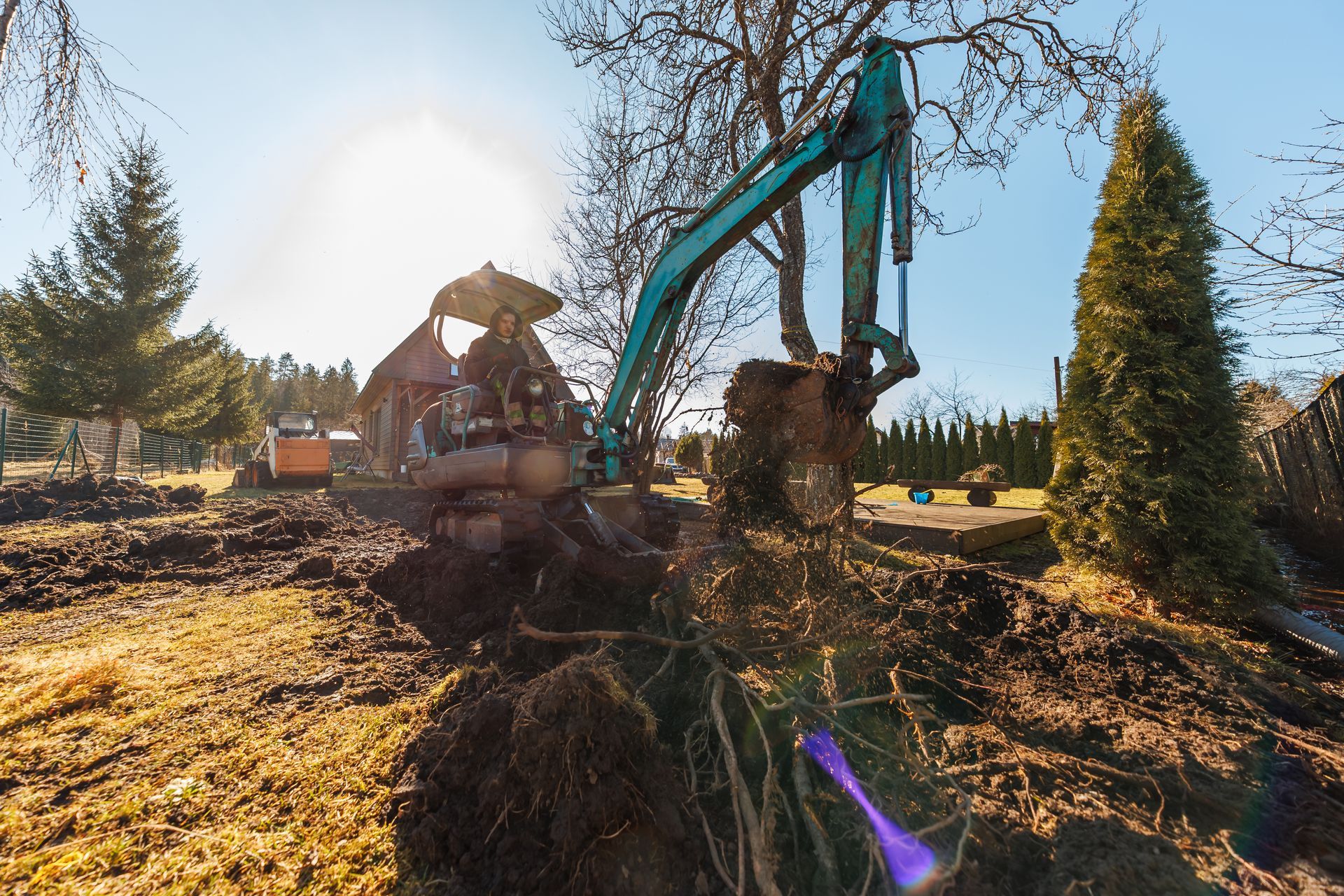 Excavator digging a trench beside a house in a sunny yard, with dirt piles and trees nearby