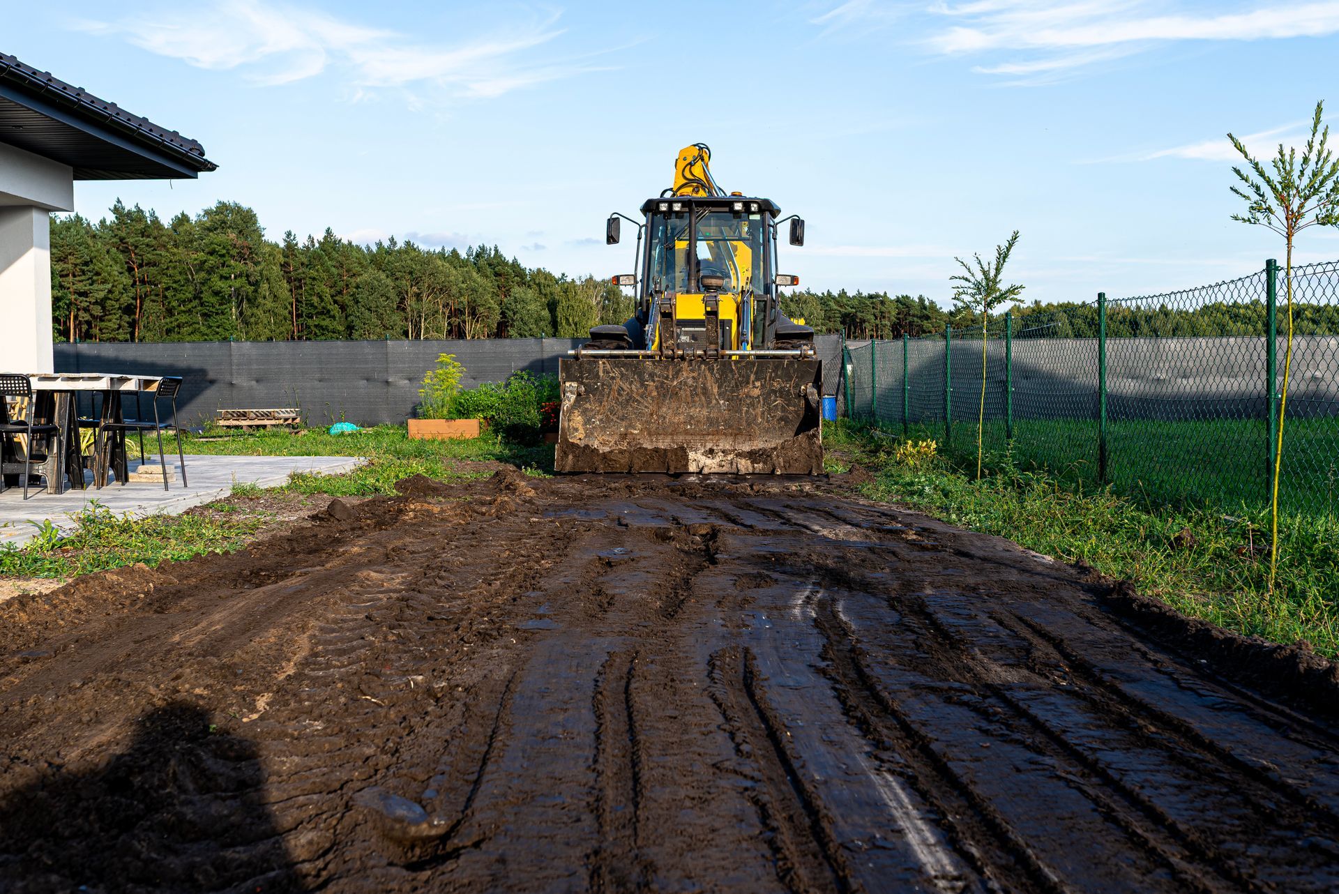 Bulldozer pushing mud on a muddy path beside a fenced yard on a sunny day