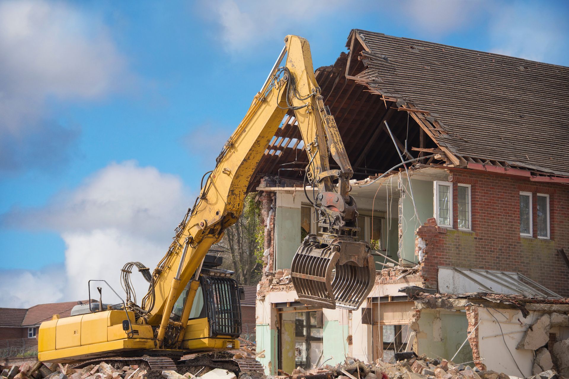 Excavator demolishing a house with debris falling beside a partly torn-down brick building Excavator demolishing a brick house with debris falling, against a blue sky.