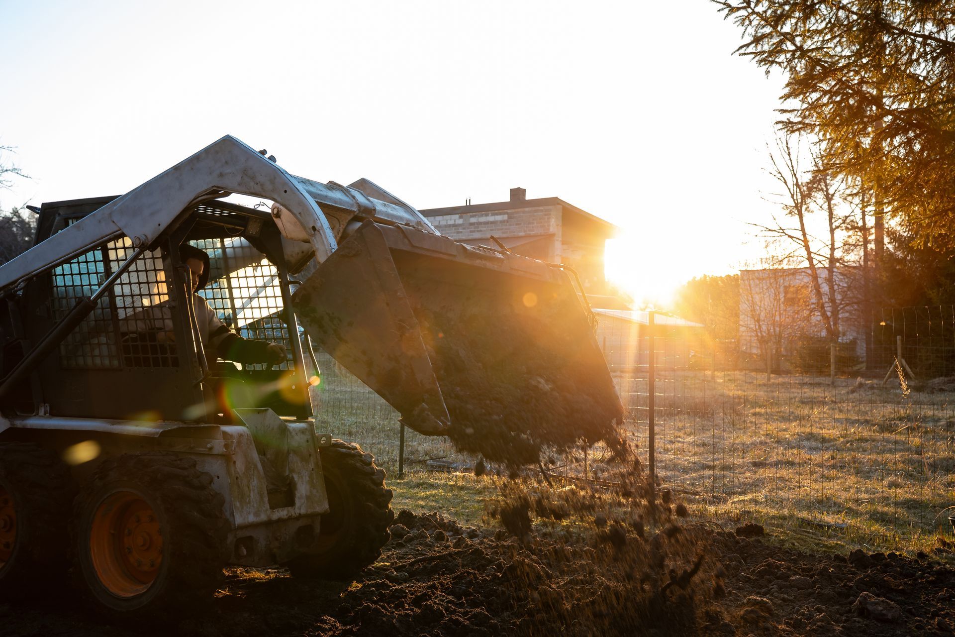 Skid steer loading rubble at sunset beside a house, with trees and bright glare in the background Skid steer loading rubble at sunset beside a house, with trees and bright glare in the background