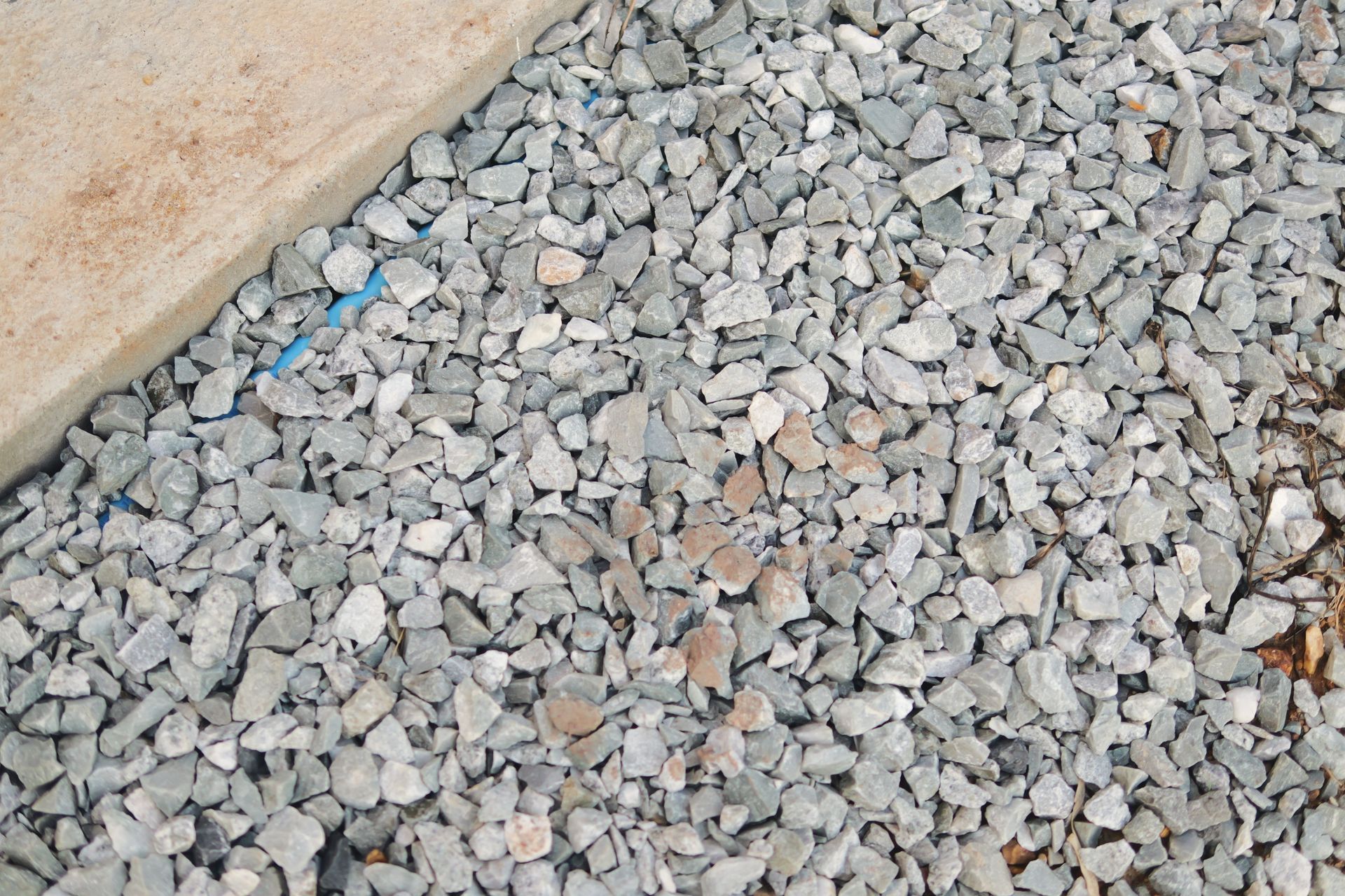 Gray gravel beside a beige wall, with a few small blue and tan stones scattered among the rocks Gray gravel beside a beige wall, with a few small blue and tan stones scattered among the rocks