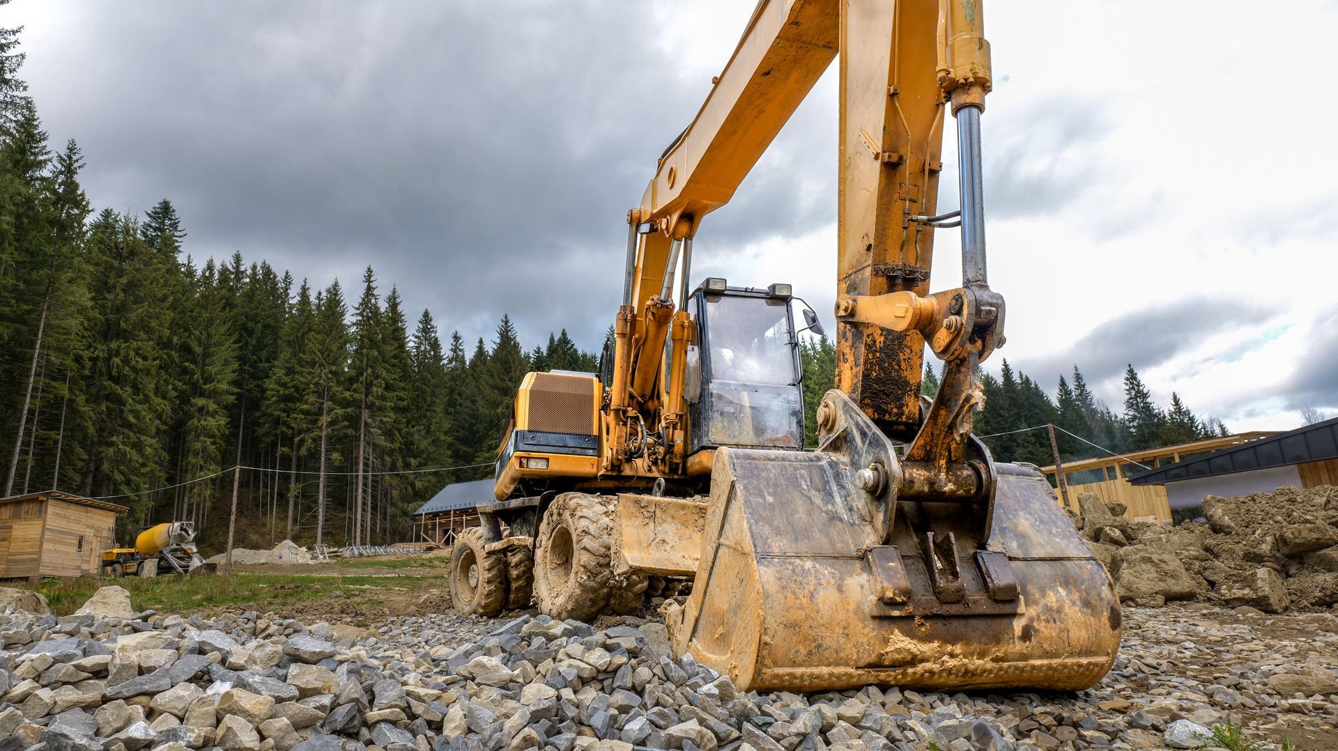 Yellow excavator digging rocks at a construction site under cloudy skies, surrounded by pine trees. Yellow excavator digging rocks at a construction site under cloudy skies, surrounded by pine trees.