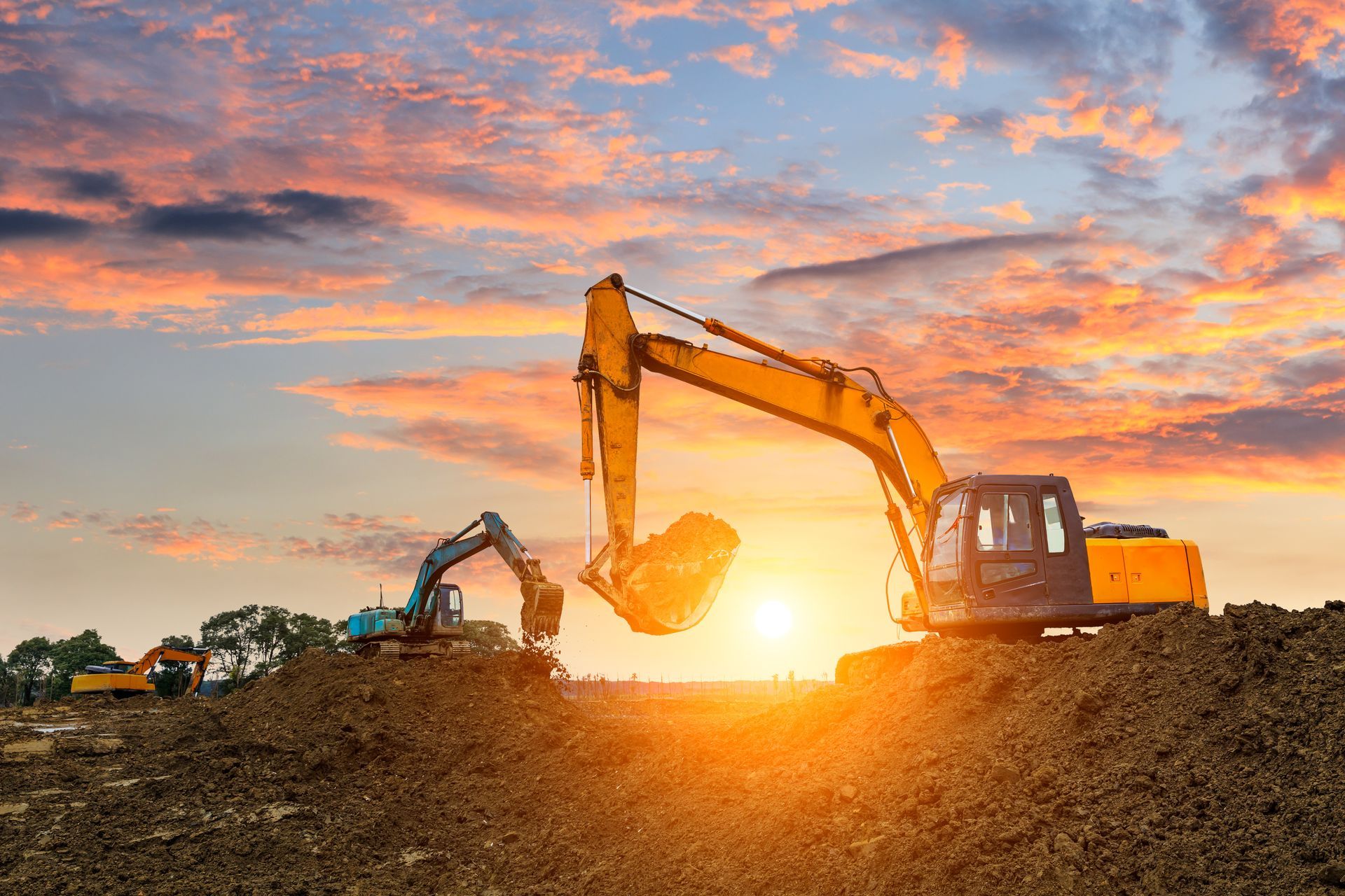 Excavators working on a dirt mound at sunset, with orange sky and bright sun on the horizon