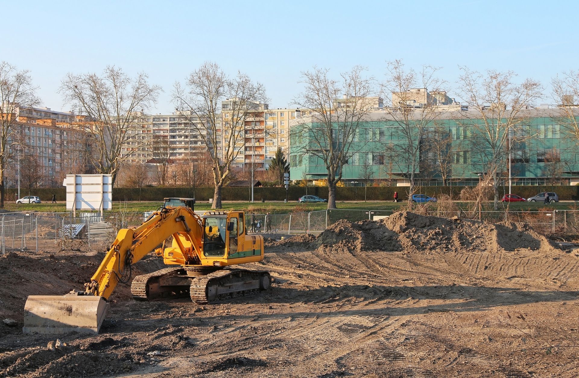 Yellow road roller and grader working on a dirt road in a rural field. Yellow excavator at a construction site with dirt piles and apartment buildings in the background