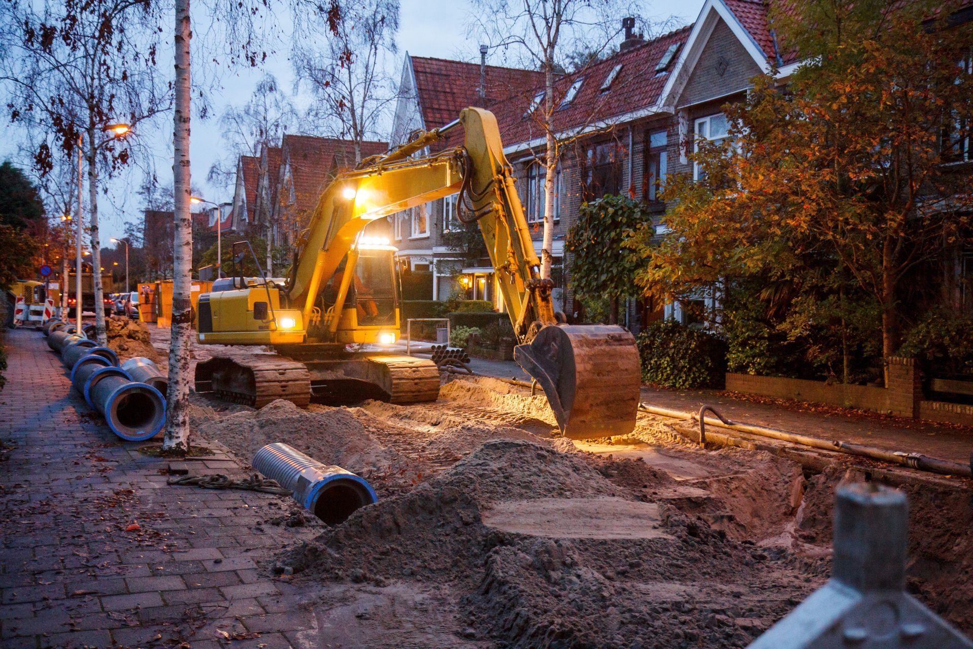 Excavator digging a muddy trench beside a tree-lined residential street at dusk Excavator digging a muddy trench beside a tree-lined residential street at dusk