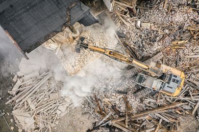 Excavator clearing debris at a demolition site, with dust rising around broken wood and rubble. Excavator clearing debris at a demolition site, with dust rising around broken wood and rubble.